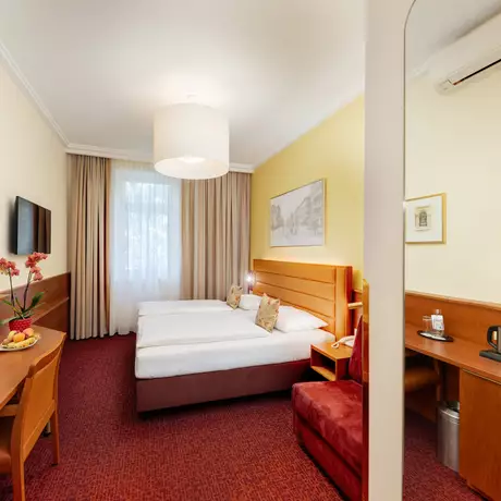 Ein gemütliches Hotelzimmer mit zwei Einzelbetten, einem großen Fenster mit Vorhängen und einem Kunstwerk an der Wand. Ein Schreibtisch mit Stuhl, Obstschale und einem Wasserkocher sind ebenfalls vorhanden. Der Boden ist mit rotem Teppich ausgelegt und die Wände sind in sanften Farben gehalten.