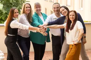 Sechs Personen stehen in einer Gruppe im Freien und legen ihre Hände übereinander. Sie lächeln freundlich und scheinen eine positive Atmosphäre zu genießen. Die Umgebung ist hell und freundlich gestaltet, mit einem klaren Hintergrund.