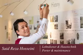 Ein junger Mann mit dunklen Haaren und einem freundlichen Lächeln hängt ein Bild an einer Wand. Im Hintergrund sind historische Fotos und Informationen zu sehen. Der Text am unteren Rand nennt seinen Namen und seine Position im Bereich Hausservice und Wartung.