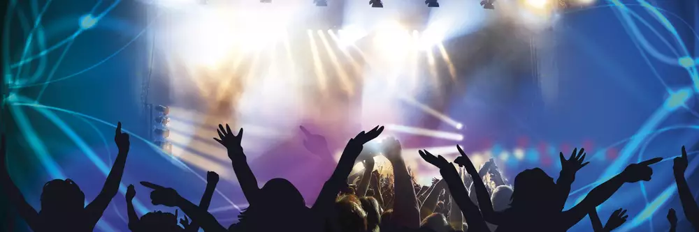 Eine Menschenmenge hebt die Hände auf einem Konzert oder einer Veranstaltung. Im Hintergrund sind helle Lichter und eine Bühne zu sehen, die eine festliche Atmosphäre erzeugen.