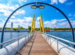 Eine Holzbrücke mit weißen Geländern führt über Wasser. Gelbe Stützpfeiler sind in einer Bogenform angeordnet. Im Hintergrund sind grünes Ufer, Gebäude und ein Leuchtturm sichtbar. Der Himmel ist blau mit einigen Wolken.