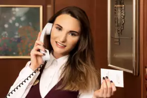 Eine Frau mit langen, braunen Haaren spricht am Telefon und hält eine Visitenkarte in der Hand. Sie trägt eine hellblau-weiße Bluse und eine dunkle Weste. Im Hintergrund sind dekorative Details wie ein Bild und ein Plüschbär zu sehen.