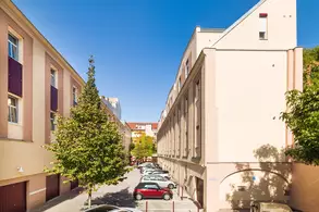 Eine Straße zwischen zwei Gebäuden mit Autos, die auf einem Parkplatz geparkt sind. Auf der linken Seite steht ein Baum. Der Himmel ist klar und blau. Diffuse Tageslichtverhältnisse.