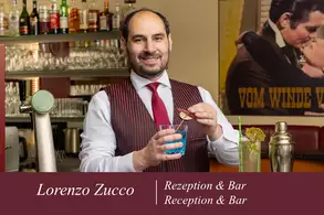 Ein Barkeeper namens Lorenzo Zucco lächelt und hält ein Glas mit blauer Flüssigkeit in der Hand. Im Hintergrund sind alkoholische Getränke und eine dekorative Bar zu sehen.