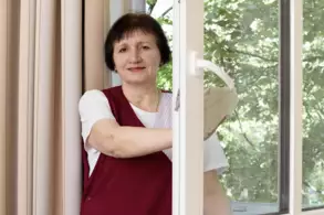 Eine Frau mit kurzen, dunklen Haaren steht am offenen Fenster und hält einen Fenstergriff. Sie trägt ein weißes T-Shirt und eine rote Schürze. Im Hintergrund sind grüne Bäume zu sehen.