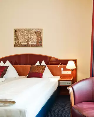Das Bild zeigt ein Hotelzimmer mit einem Doppelbett, einem Nachttisch mit Lampe und einem roten Sessel. An der Wand hängt ein dekoratives Kunstwerk. Die Vorhänge sind rot gestreift, und es gibt Tageslicht, das durch ein Fenster fällt.