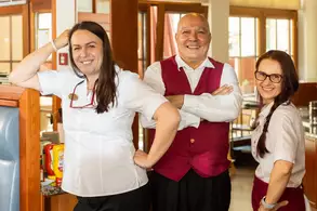 Drei Mitarbeiter stehen in einem Restaurant zusammen. Die Frau links trägt ein weißes Hemd und hat die Hand an der Hüfte. Der Mann in der Mitte trägt eine Weste und lächelt. Die Frau rechts hat ein rotes Oberteil und eine Brille. Sie wirken freundlich und lässig.