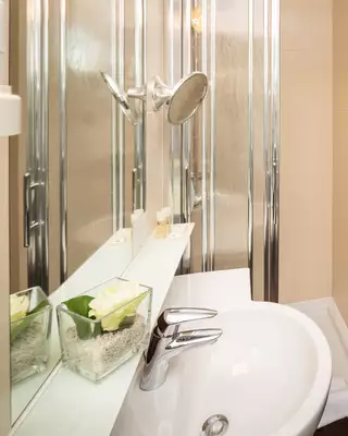 Das Bild zeigt ein modernes Badezimmer mit einer Dusche, einem Waschbecken und einem Spiegel. Im Hintergrund ist ein angrenzendes Zimmer mit einem Bett und einem Schreibtisch sichtbar. Die Einrichtung ist einfach und hell.