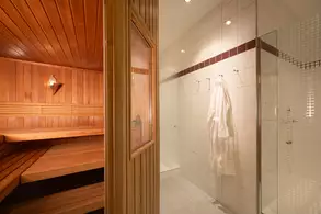 Ein Blick in einen Wellnessbereich mit einer Holzgarten-Sauna auf der linken Seite und einem modernen Duschbereich auf der rechten Seite. Ein weißer Bademantel hängt an einer Wandhalterung. Der Raum ist hell erleuchtet und vermittelt ein entspannendes Ambiente.