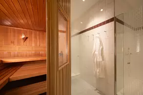 Ein Blick in einen Wellnessbereich mit einer Holzgarten-Sauna auf der linken Seite und einem modernen Duschbereich auf der rechten Seite. Ein weißer Bademantel hängt an einer Wandhalterung. Der Raum ist hell erleuchtet und vermittelt ein entspannendes Ambiente.