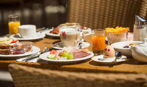 Ein gedeckter Tisch mit Kaffee, Gebäck und Obstschale unter einem Sonnenschirm im Freien.