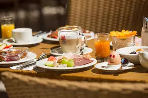 Ein gedeckter Tisch mit Kaffee, Gebäck und Obstschale unter einem Sonnenschirm im Freien.