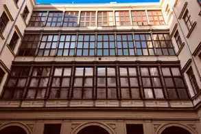 Das Bild zeigt die Fassade eines Gebäudes mit mehreren großen Fenstern und einem klaren Himmel im Hintergrund. Die Fenster sind in einem modernen Stil angeordnet, während die architektonischen Details an der unteren Gebäudeteile nicht näher erkennbar sind.