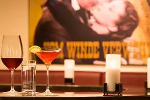 Two cocktails are on a bar: a glass of red wine, a martini with a lemon twist, and a tall glass of water. The background features a vintage-style poster, adding a stylish touch to the scene. Soft candlelight enhances the ambiance.