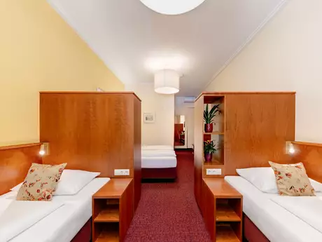 Zwei Einzelbetten in einem hellen Innenraum, getrennt durch Holzwände mit Nachttischlampe und Pflanzen. Im Hintergrund befindet sich ein weiteres Bett. Die Wände sind in sanften Farben gehalten, der Boden ist mit Teppich ausgelegt.