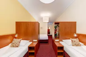 Zwei Einzelbetten in einem hellen Innenraum, getrennt durch Holzwände mit Nachttischlampe und Pflanzen. Im Hintergrund befindet sich ein weiteres Bett. Die Wände sind in sanften Farben gehalten, der Boden ist mit Teppich ausgelegt.