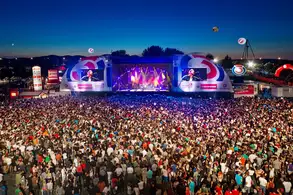 Eine große Menschenmenge versammelt sich vor einer Bühne bei einem Konzert oder Festival. Die Bühne ist beleuchtet und zeigt eine Performance, während der Himmel bei Sonnenuntergang leuchtet. Umgebende Strukturen und Veranstaltungselemente sind sichtbar.