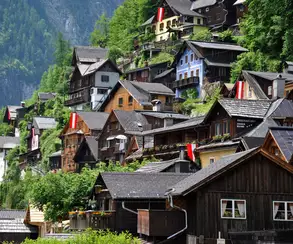 Das Bild zeigt eine malerische Berglandschaft mit traditionellen, bunten Häusern, die an einem Hang gebaut sind. Einige Häuser sind mit roten und weißen Fahnen geschmückt. Umgebende Bäume und grünes Gelände tragen zur idyllischen Atmosphäre bei.