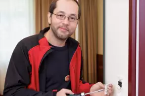 Ein Mann mit Brille und einem roten und schwarzen Oberteil hält einen Schraubendreher in der Hand. Er arbeitet an einer Steckdose, die an einer Wand montiert ist. Im Hintergrund sind Vorhänge sichtbar.
