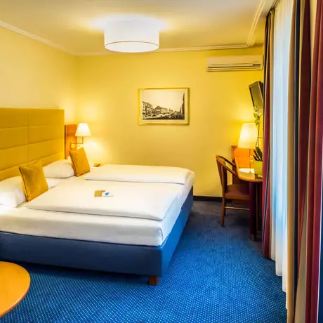 Ein helles Hotelzimmer mit zwei Einzelbetten, Nachttischen und Lampen. An der Wand hängt ein Bild. In einer Ecke steht ein Schreibtisch mit einem Stuhl. Ein kleiner Tisch mit einer Obstschale ist im Vordergrund sichtbar. Der Boden ist blau und die Vorhänge sind gestreift.