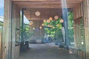 Eingang zu einem Gebäude mit holzverkleideter Fassade und der Aufschrift "Herzlich Willkommen". Hell erleuchtet mit hängenden Lampen, umgeben von Pflanzen. Jemand geht im Hintergrund.