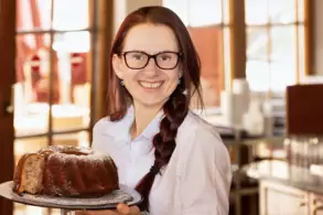 Eine lächelnde Frau mit Brille trägt einen großen Kuchen auf einem Teller. Sie steht in einer hellen, geschäftigen Umgebung mit Fenstern im Hintergrund.