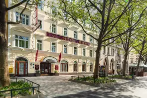 Fassade des Hotel Wien mit gelbem Stuck, rotem Vordach und großen Bäumen im Vordergrund.