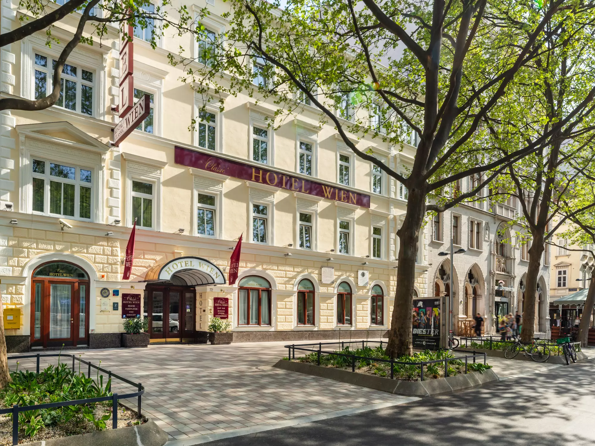 Fassade des Hotel Wien mit gelbem Stuck, rotem Vordach und großen Bäumen im Vordergrund.