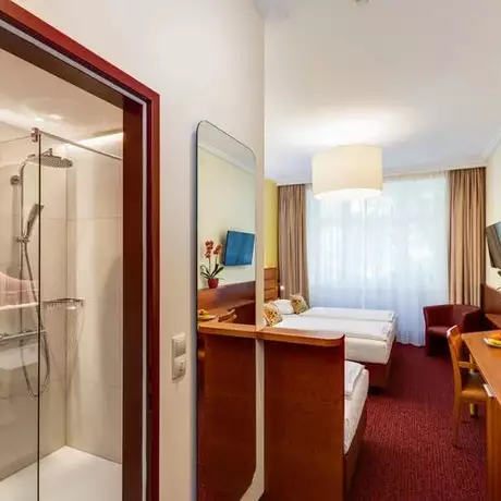 Ein helles Hotelzimmer mit einem Doppelbett und einem Einzelbett, einem Schreibtisch, einem Sessel und einem Fernseher. Auf der linken Seite befindet sich ein modernes Badezimmer mit einer Duschkabine. Natürliches Licht fällt durch ein Fenster, das mit Vorhängen dekoriert ist.