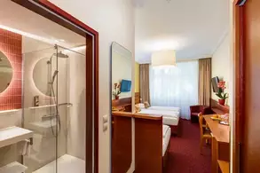 Ein helles Hotelzimmer mit einem Doppelbett und einem Einzelbett, einem Schreibtisch, einem Sessel und einem Fernseher. Auf der linken Seite befindet sich ein modernes Badezimmer mit einer Duschkabine. Natürliches Licht fällt durch ein Fenster, das mit Vorhängen dekoriert ist.