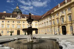 Der Innenhof eines barocken Gebäudes mit einem Brunnen in der Mitte. Der Brunnen zeigt eine Statue und ist von einer Steinumrandung umgeben. Im Hintergrund sind die gelben Fassaden des Gebäudes und ein blauer Himmel mit vereinzelten Wolken zu sehen.