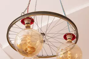 Eine ungewöhnliche Deckenleuchte, bestehend aus einem Rad mit drei großen, runden Glühbirnen. Die Glühbirnen sind transparent und leuchten warm. Das Rad ist an Ketten und bunten Kabeln befestigt, die von der Decke hängen. Der Hintergrund ist schlicht und hell.