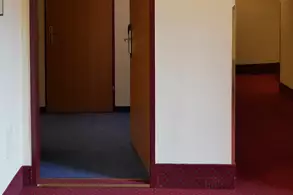 Ein Flur mit einer offenen Tür, durch die ein Badezimmer sichtbar ist. An der Wand neben der Tür hängt ein Namensschild mit der Zimmernummer. Der Boden ist mit Teppich ausgelegt und die Wände sind hell gefärbt.