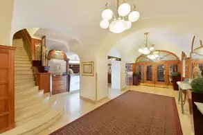 Das Bild zeigt die Lobby eines Hotels mit einer eleganten Treppe, die nach oben führt. Die Wände sind hell gestrichen und eine Teppichfläche sorgt für einen gemütlichen Eindruck. Große, runde Kronleuchter hängen von der Decke und beleuchten den Raum.