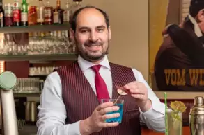 Ein Barkeeper in einem gestreiften Weste lächelt und hält ein Glas mit einem blauen Getränk. Er steht an einer Bar mit vielen Spirituosen im Hintergrund.