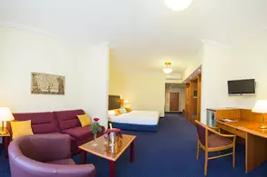 Ein Hotelzimmer mit einem Sitzbereich, bestehend aus einem roten Sofa und einem Stuhl. Ein Tisch steht in der Mitte, darauf eine Flasche und Glas. Im Hintergrund befindet sich ein Bett und ein Schreibtisch mit einem Fernseher. Die Wände sind hell und es gibt mehrere Lichtquellen.