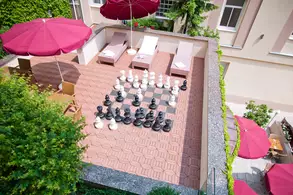 An aerial view of a rooftop terrace featuring a large outdoor chessboard with black and white pieces arranged on it. Surrounding the chessboard are lounge chairs and two red umbrellas providing shade, along with greenery in the background.