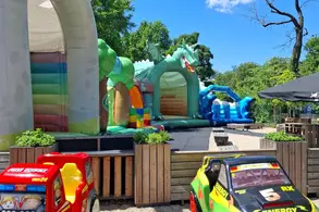 Bunte Hüpfburgen in einer Freizeitgestaltung sind sichtbar, darunter ein Einhorn und ein Drache. Zwei Spielzeugautos stehen im Vordergrund, während blauer Himmel und Bäume die Szene umrahmen.