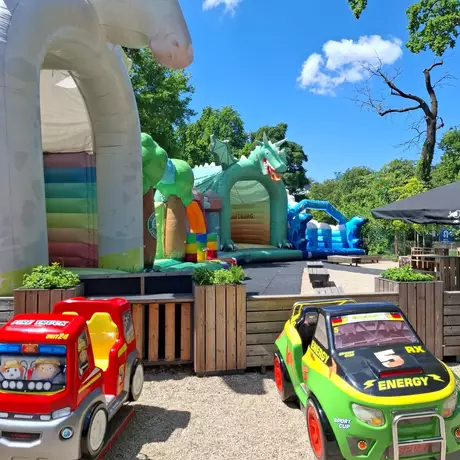 Bunte Hüpfburgen in einer Freizeitgestaltung sind sichtbar, darunter ein Einhorn und ein Drache. Zwei Spielzeugautos stehen im Vordergrund, während blauer Himmel und Bäume die Szene umrahmen.