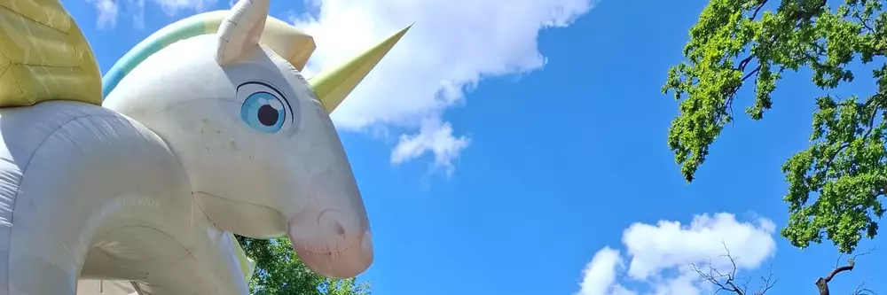 Bunte Hüpfburgen in einer Freizeitgestaltung sind sichtbar, darunter ein Einhorn und ein Drache. Zwei Spielzeugautos stehen im Vordergrund, während blauer Himmel und Bäume die Szene umrahmen.