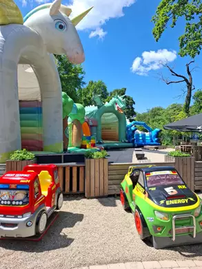 Bunte Hüpfburgen in einer Freizeitgestaltung sind sichtbar, darunter ein Einhorn und ein Drache. Zwei Spielzeugautos stehen im Vordergrund, während blauer Himmel und Bäume die Szene umrahmen.