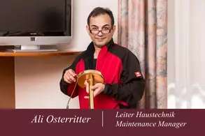 Ein Mann in Arbeitskleidung hält ein Werkzeug in der Hand. Im Hintergrund sind ein Fernseher und Vorhänge zu sehen. Auf einem Banner sind sein Name und seine Berufsbezeichnung als Leiter Haustechnik abgebildet.