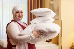 Eine Frau trägt vier weiße Kissen in ihren Armen. Sie steht in einem hellen Zimmer mit Vorhängen im Hintergrund. Das Bild vermittelt eine freundliche und einladende Atmosphäre.