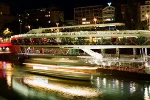 Eine moderne Aussichtsplattform über einem beleuchteten Gewässer in der Nacht. im Vordergrund fährt ein Schiff mit Lichtspuren vorbei. Im Hintergrund sind Gebäude und Straßenlaternen sichtbar.