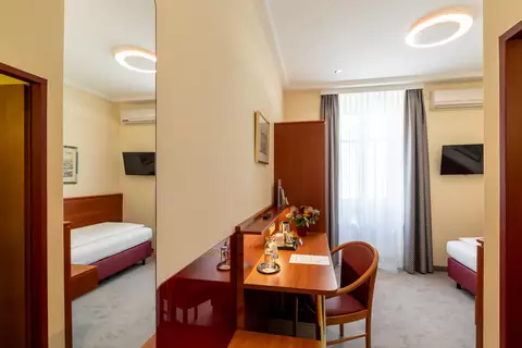 Ein helles Zimmer mit einem Einzelbett, einem Schreibtisch und einem Stuhl. Am Schreibtisch stehen eine Vase mit Blumen und eine Taschenlampe. Ein großes Fenster mit Vorhang lässt viel Tageslicht herein. Im Hintergrund sind Wände und ein Fernseher sichtbar.