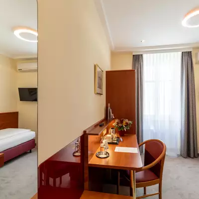 Ein helles Zimmer mit einem Einzelbett, einem Schreibtisch und einem Stuhl. Am Schreibtisch stehen eine Vase mit Blumen und eine Taschenlampe. Ein großes Fenster mit Vorhang lässt viel Tageslicht herein. Im Hintergrund sind Wände und ein Fernseher sichtbar.