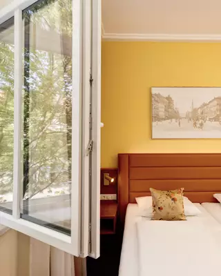 Ein helles Hotelzimmer mit einem Fenster, das weit geöffnet ist. Im Raum befinden sich zwei Betten mit weißen Bettwäsche und dekorativen Kissen. An der Wand hängt ein Bild einer urbanen Szenerie. Die Wand ist in einem auffälligen Gelbton gestrichen.