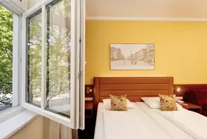 Ein helles Hotelzimmer mit einem Fenster, das weit geöffnet ist. Im Raum befinden sich zwei Betten mit weißen Bettwäsche und dekorativen Kissen. An der Wand hängt ein Bild einer urbanen Szenerie. Die Wand ist in einem auffälligen Gelbton gestrichen.