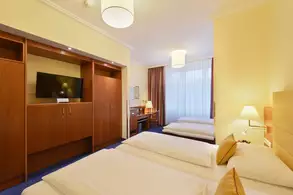 Ein helles Zimmer mit zwei Doppelbetten, einem Schreibtisch und einem Fernseher an der Wand. Ein Fenster mit Vorhängen lässt Tageslicht herein. Es gibt eine Holzkommode und eine Lampe auf einem Nachttisch.