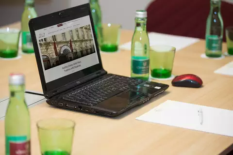Ein offenstehender Laptop auf einem Tisch zeigt eine Webseite eines Hotels. Um den Laptop herum stehen grüne Gläser und Flaschen mit Wasser. Daneben liegen ein Notizblock und ein Stift.